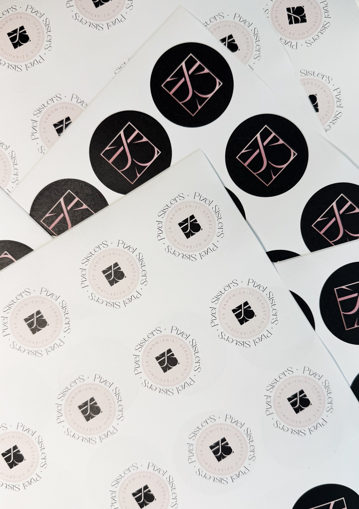 Arkusz z okrągłymi naklejkami firmowymi Pixel Sisters, z logo w dwóch wersjach kolorystycznych: czarno-różowej i czarno-białej. Naklejki ułożone pod kątem.