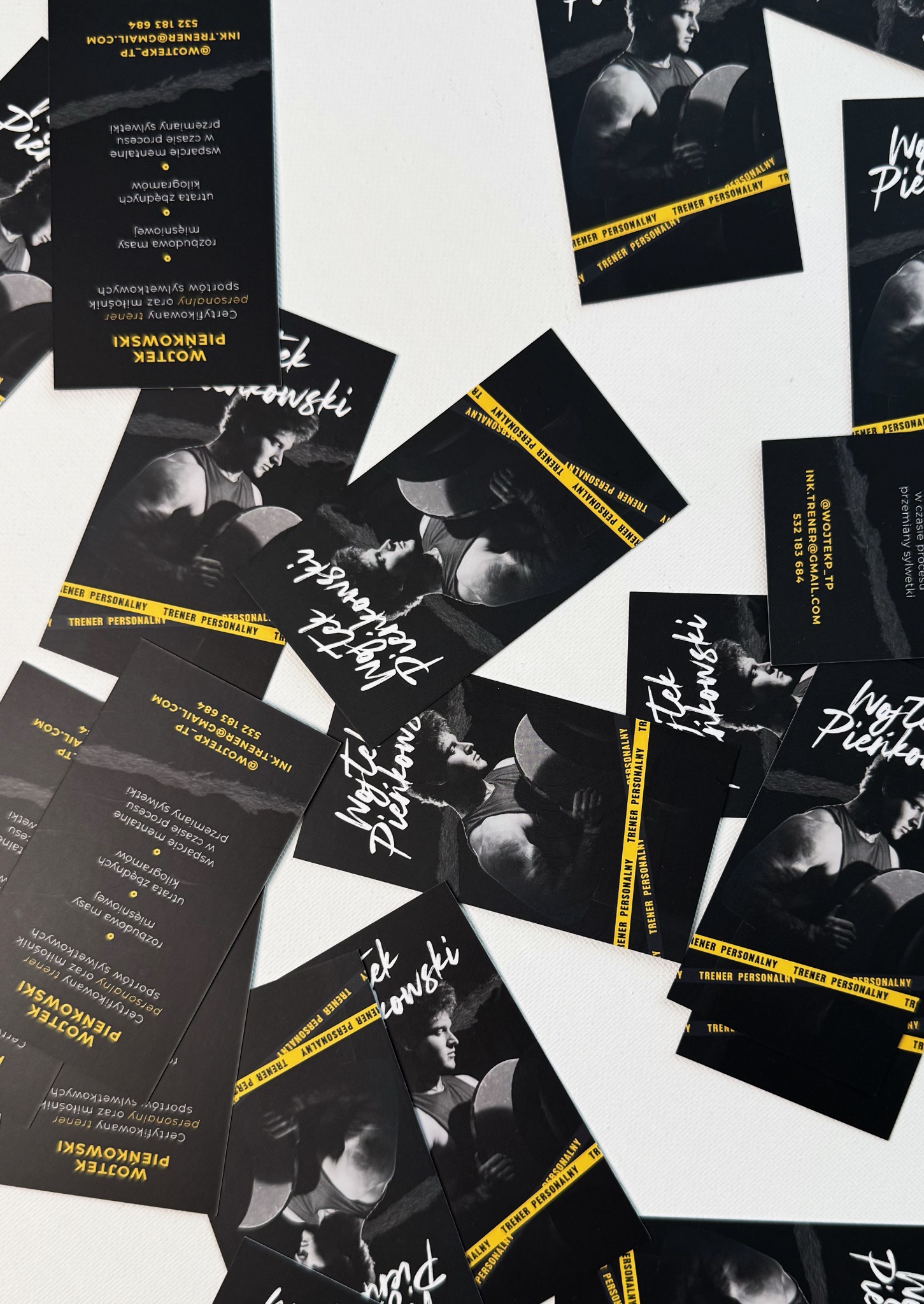 Rozrzucone wizytówki trenera personalnego w czerni i złocie, z jego zdjęciem podczas ćwiczeń i danymi kontaktowymi, na białym tle. Układ nieregularny, bliski kadr.