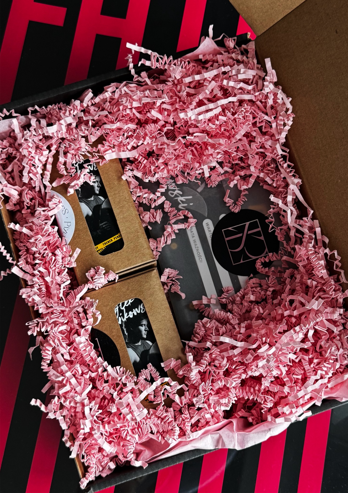 Otwarta paczka z wizytówkami w brązowych pudełkach, wypełniona różowym papierem ozdobnym, na czarnym tle z czerwonymi paskami. Prezentacja gotowych materiałów.