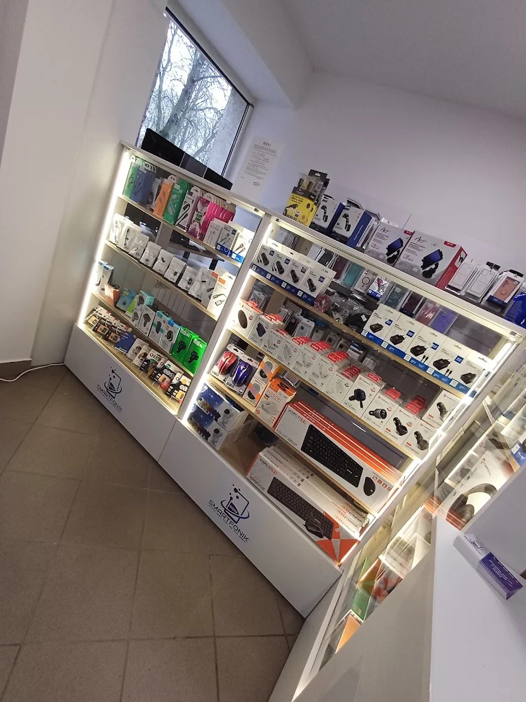 Oświetlona witryna sklepowa z akcesoriami do telefonów i komputerów, widoczne klawiatury, słuchawki i etui. Logo sklepu 'SMARTFONIK' na dolnej części witryny.