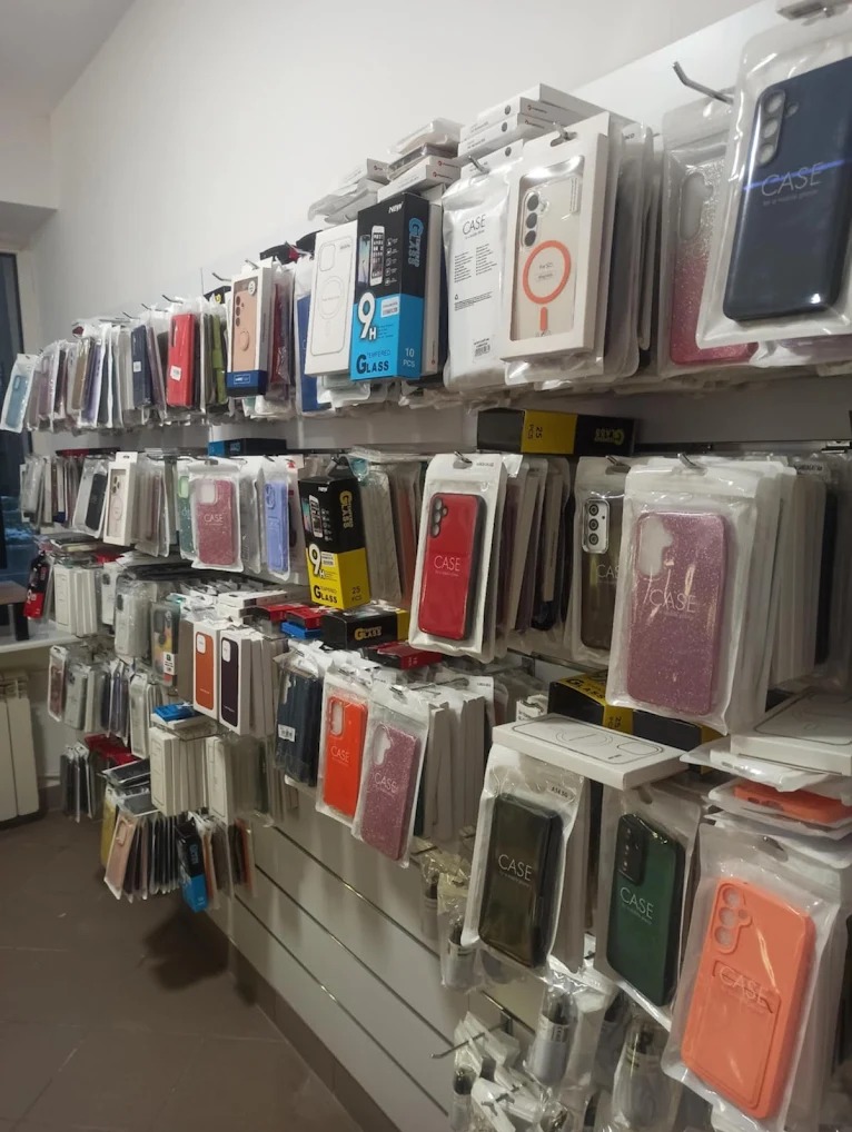 Ściana z akcesoriami do telefonów: etui w opakowaniach, szkła hartowane. Różne kolory i modele. Widok z dołu, oświetlenie sklepowe.