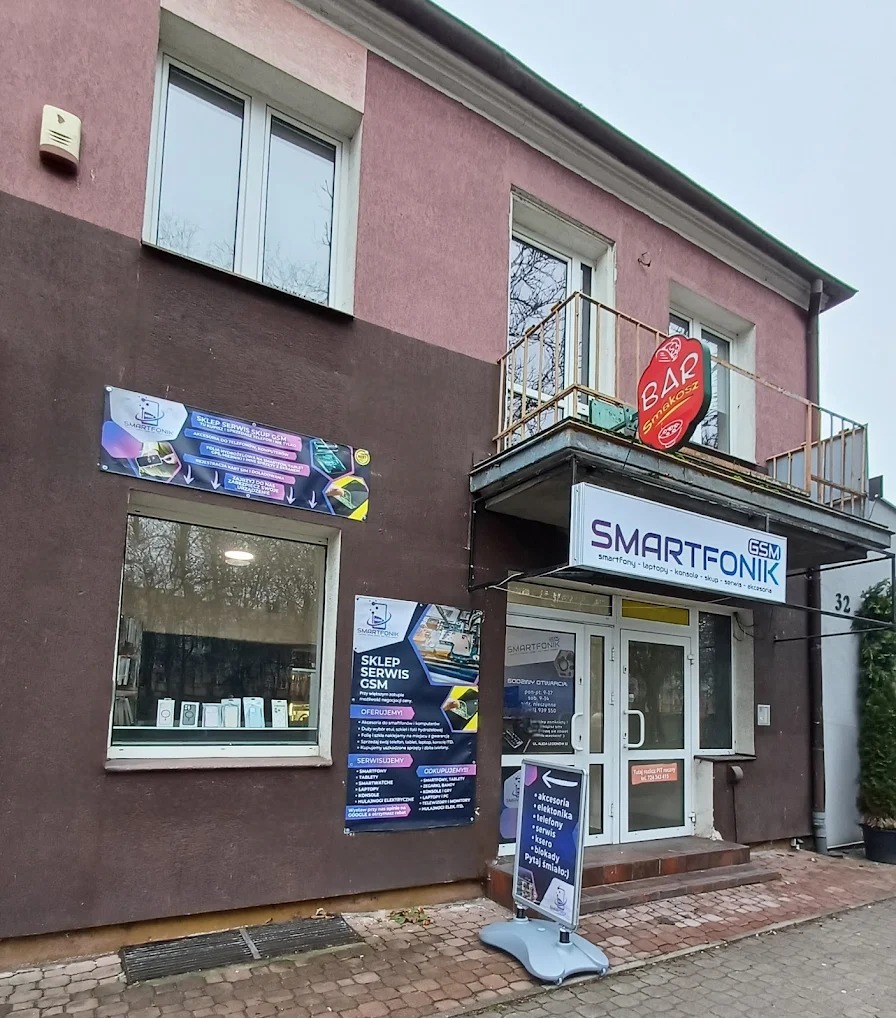 Fasada budynku z szyldem 'SMARTFONIK GSM', oferującego serwis smartfonów, laptopów i konsol. Widoczne plakaty z ofertą usług i akcesoriami. Wejście do lokalu.