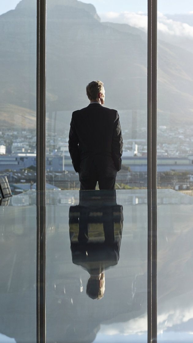 Mężczyzna w garniturze stoi tyłem, spoglądając przez okno na panoramę miasta i góry. Jego odbicie widoczne na szklanym stole.