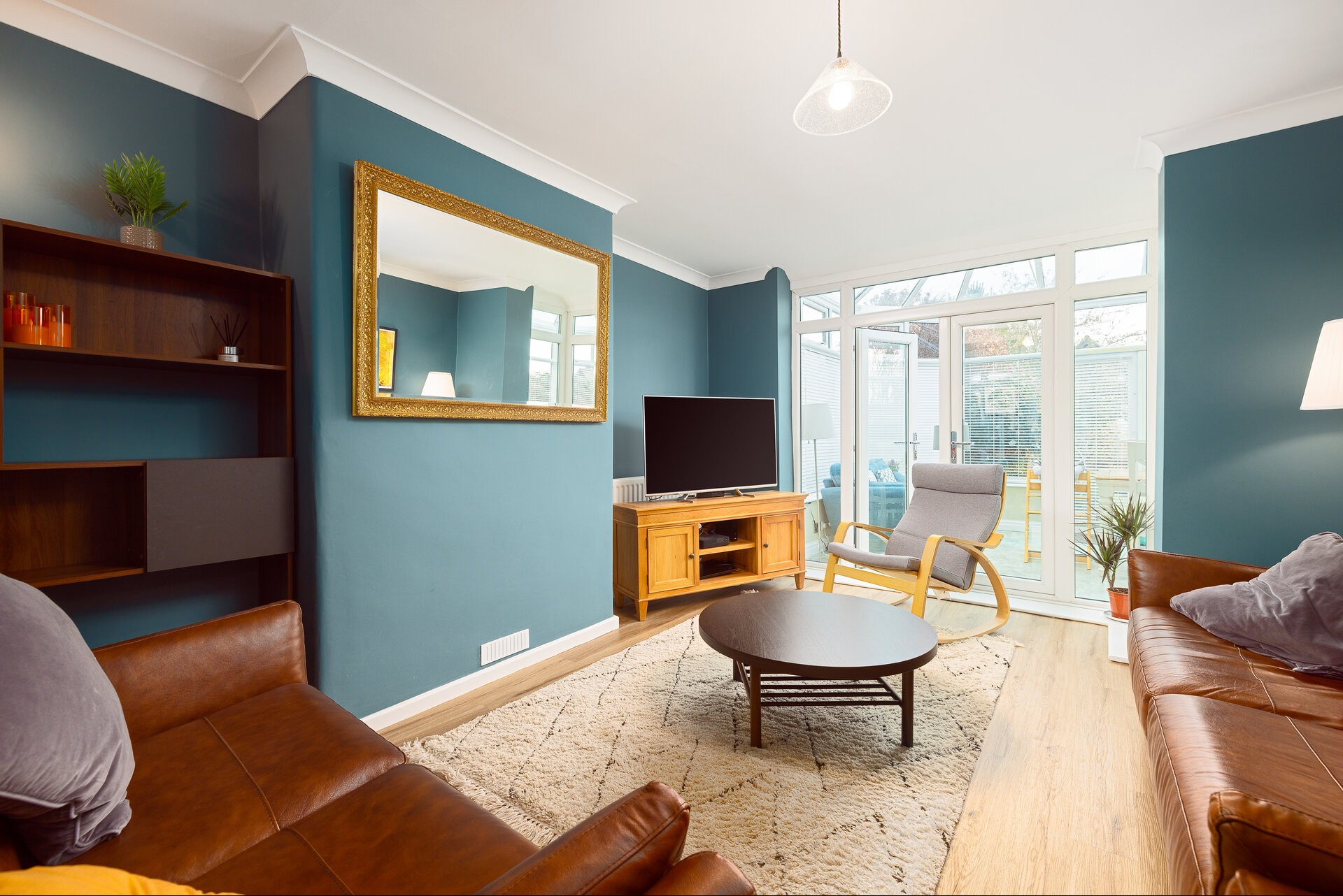 Salon w odcieniach błękitu z brązowymi skórzanymi sofami, drewnianym regałem i lustrem w złotej ramie. Widoczne okno balkonowe i dywan z geometrycznym wzorem.