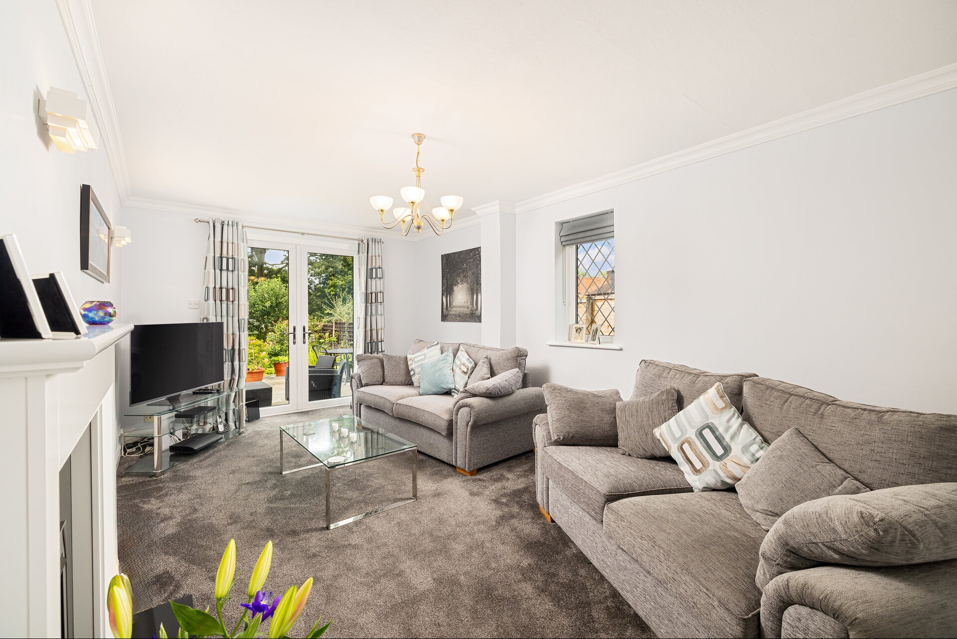 Jasne wnętrze salonu z dwoma szarymi sofami, szklanym stolikiem i telewizorem. Widoczne okno i drzwi balkonowe z widokiem na ogród. Neutralna kolorystyka, dywan w odcieniach szarości.