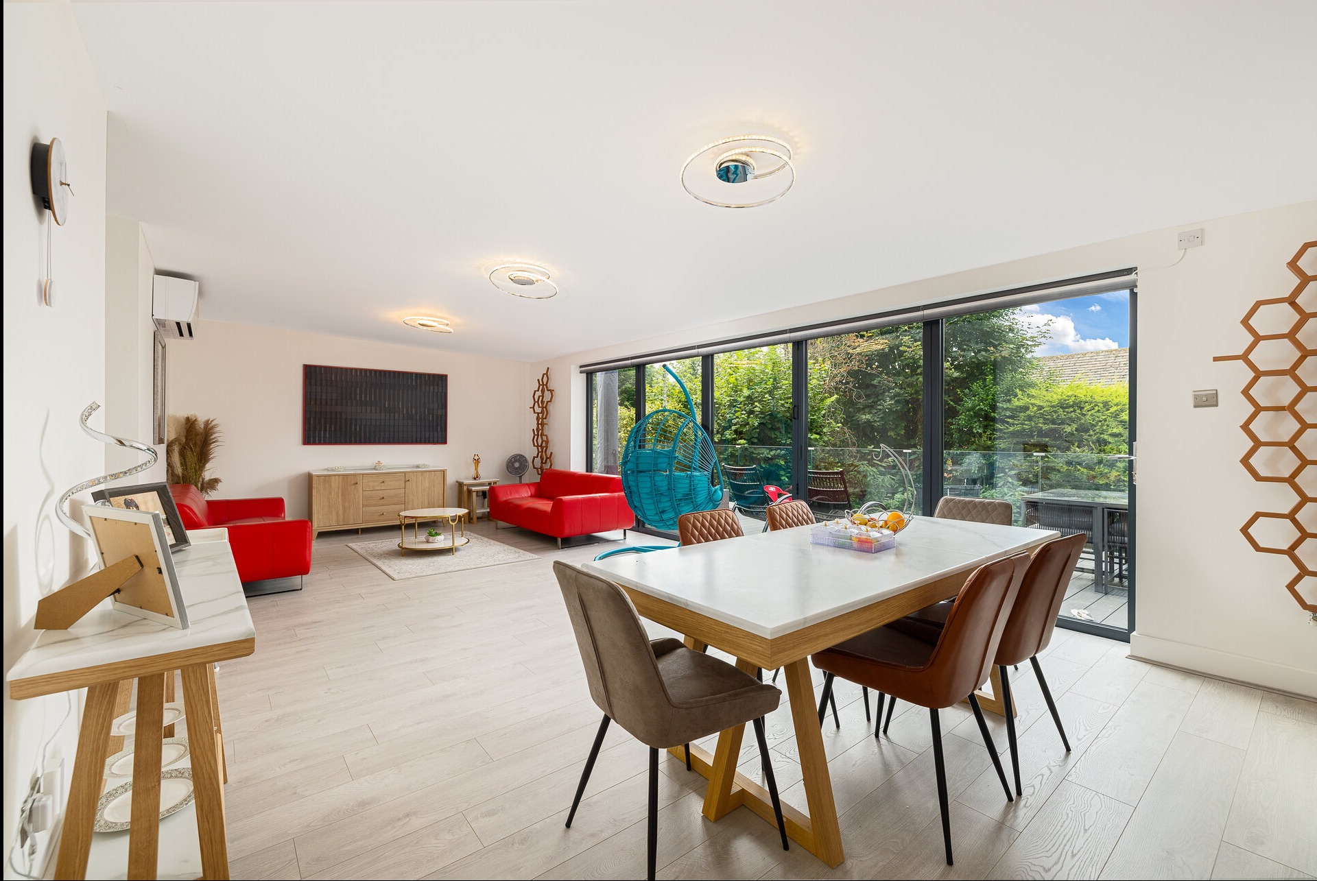 Jasne, przestronne wnętrze domu z czerwonymi sofami, stołem jadalnym i dużym oknem na ogród. Nowoczesne oświetlenie i drewniane akcenty dekoracyjne.