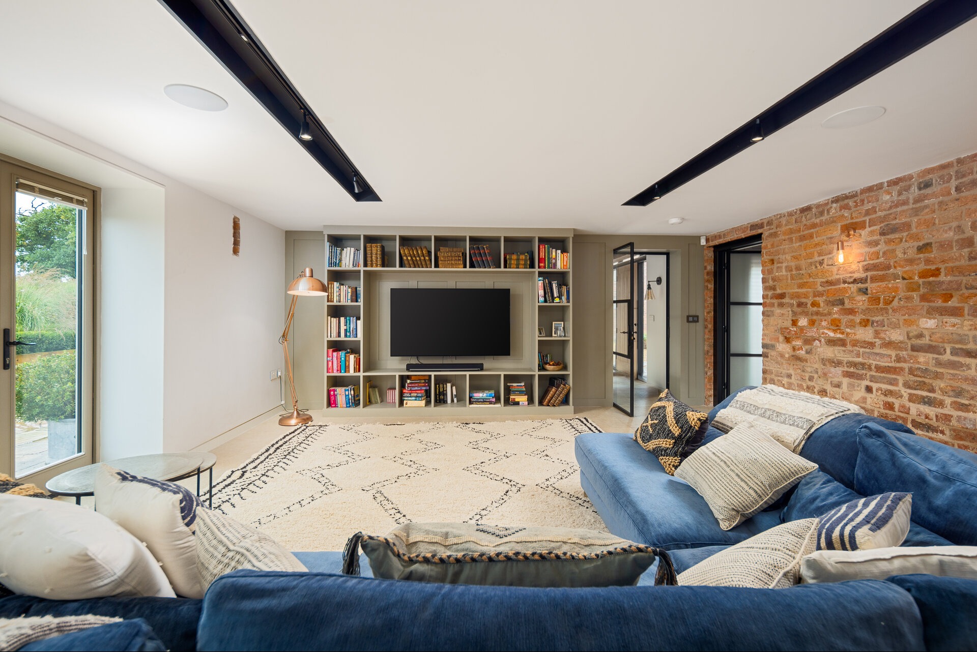 Salon z ceglaną ścianą, niebieską sofą i regałem na książki z telewizorem. Widoczne oświetlenie sufitowe i dywan w stylu boho. Duże okno z widokiem na zieleń.