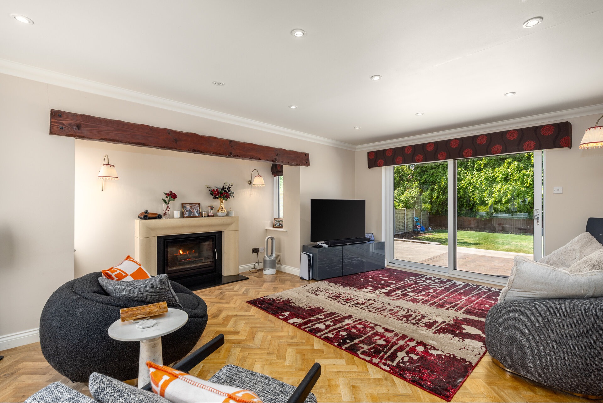 Jasny salon z kominkiem, drewnianymi belkami i dużym oknem na ogród. Parkiet w jodełkę, dywan w odcieniach czerwieni i beżu, oraz nowoczesne meble tworzą przytulną przestrzeń.