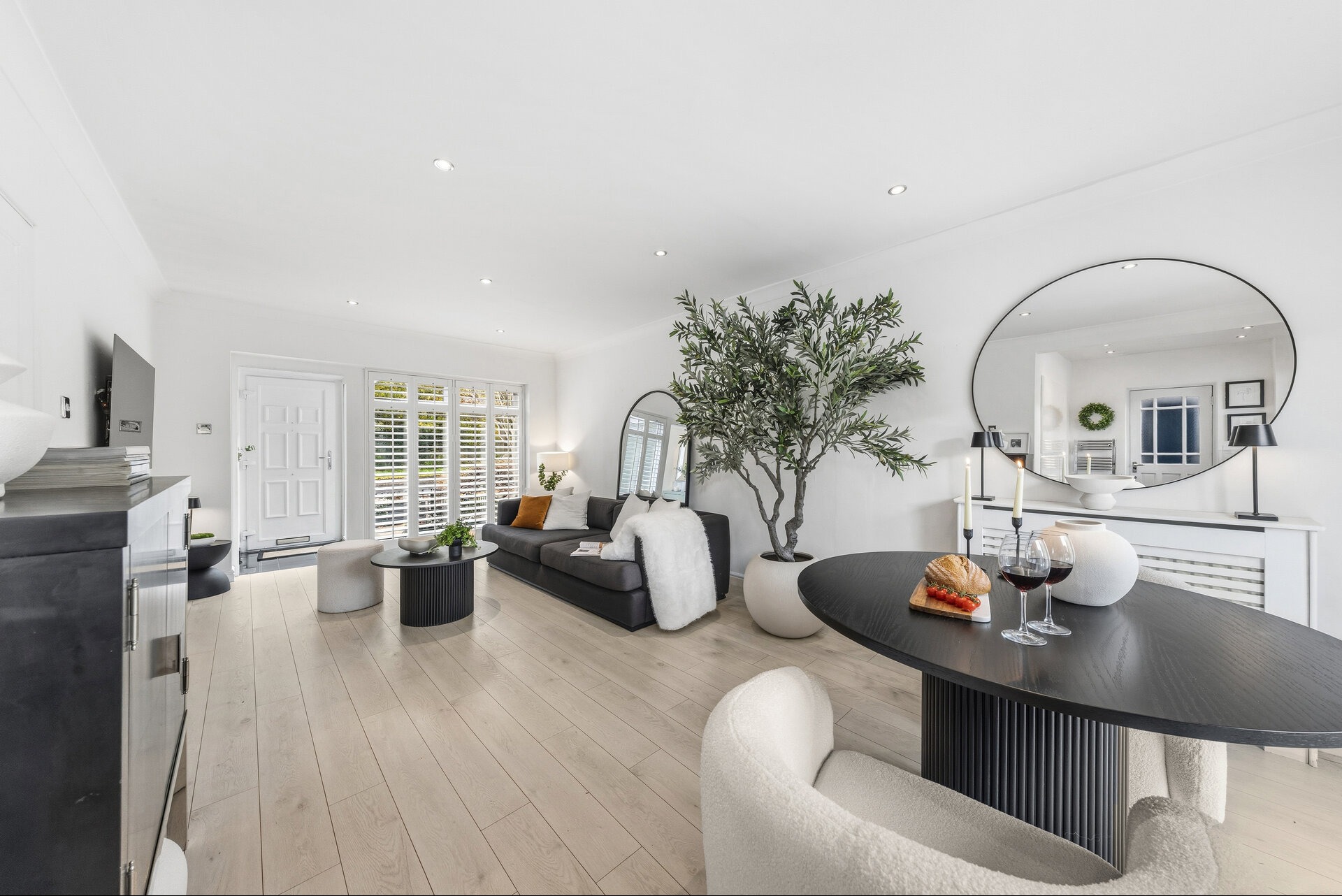 Jasne wnętrze salonu z drewnianą podłogą, szarą sofą, okrągłym stołem z przekąskami i dużym lustrem. Minimalistyczny design z rośliną w donicy.