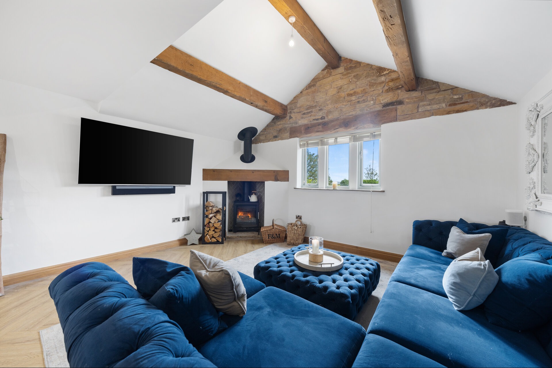 Jasne wnętrze salonu z niebieską sofą modułową, kominkiem i telewizorem. Widoczne drewniane belki stropowe i kamienna ściana. Przytulna aranżacja z elementami dekoracyjnymi.