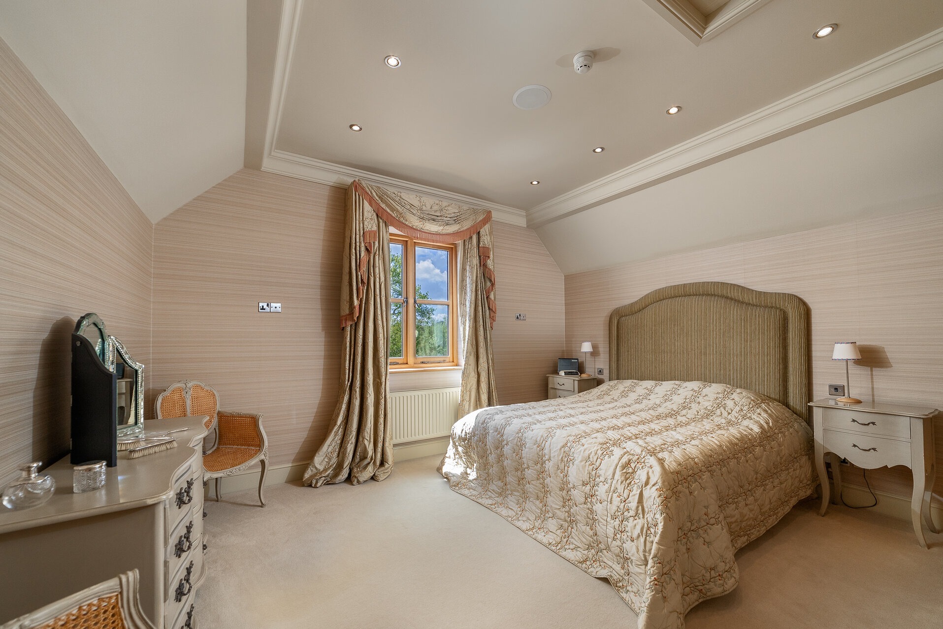 Elegancka sypialnia w beżach: łóżko z pikowaną narzutą, toaletka z lustrem, okno z dekoracyjnymi zasłonami. Subtelne oświetlenie punktowe podkreśla detale.