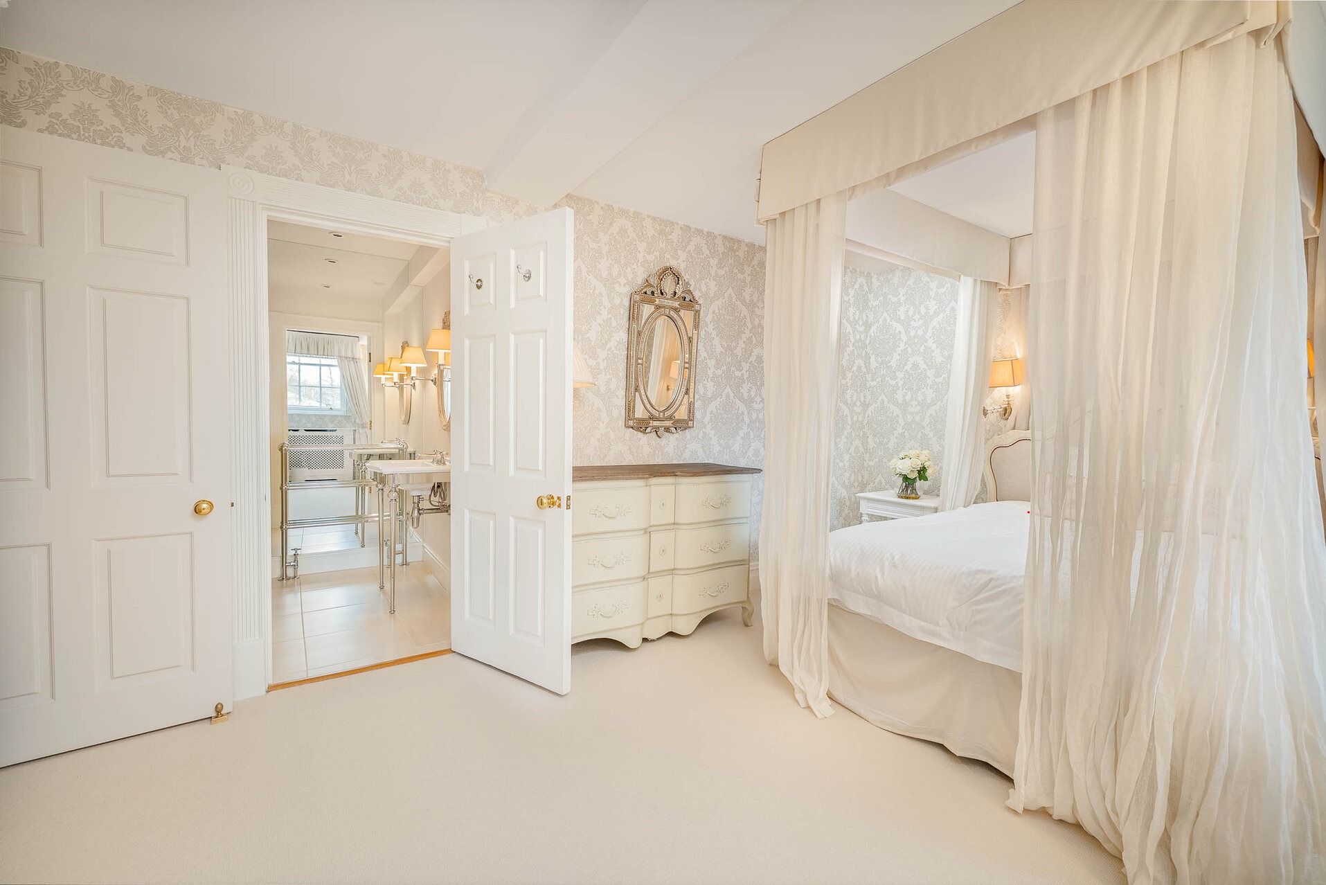 Jasne wnętrze sypialni z łazienką w tle. Baldachim nad łóżkiem, wzorzysta tapeta i kremowa komoda tworzą elegancki, spójny wystrój. Otwarta przestrzeń.