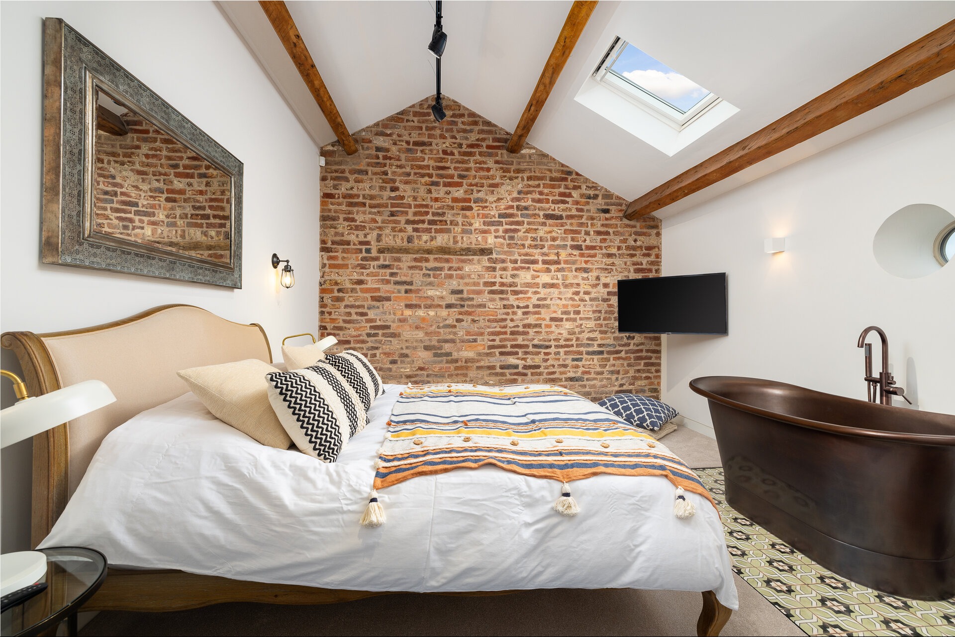 Stylowa sypialnia na poddaszu z ceglaną ścianą, drewnianymi belkami, miedzianą wanną i lustrem w ozdobnej ramie. Widoczne łóżko z dekoracyjną narzutą i poduszkami.