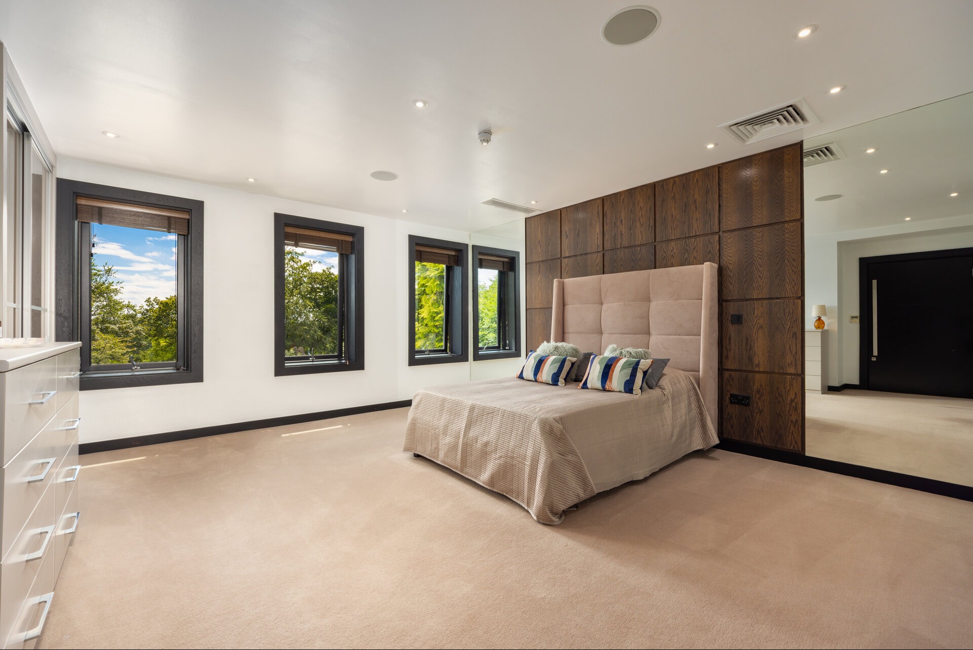 Jasna sypialnia z dużym łóżkiem, drewnianą ścianą i lustrem. Trzy okna z widokiem na zieleń. Minimalistyczny design z elementami drewna i beżu.