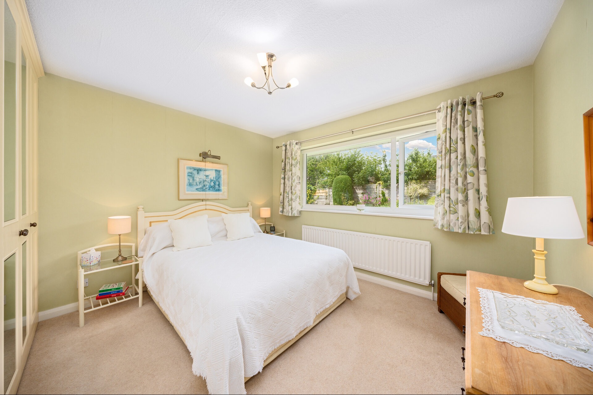 Jasna sypialnia z białym łóżkiem, kremowymi ścianami i oknem z widokiem na zieleń. Na ścianie obrazek w ramce, a obok łóżka szafka nocna z lampką.