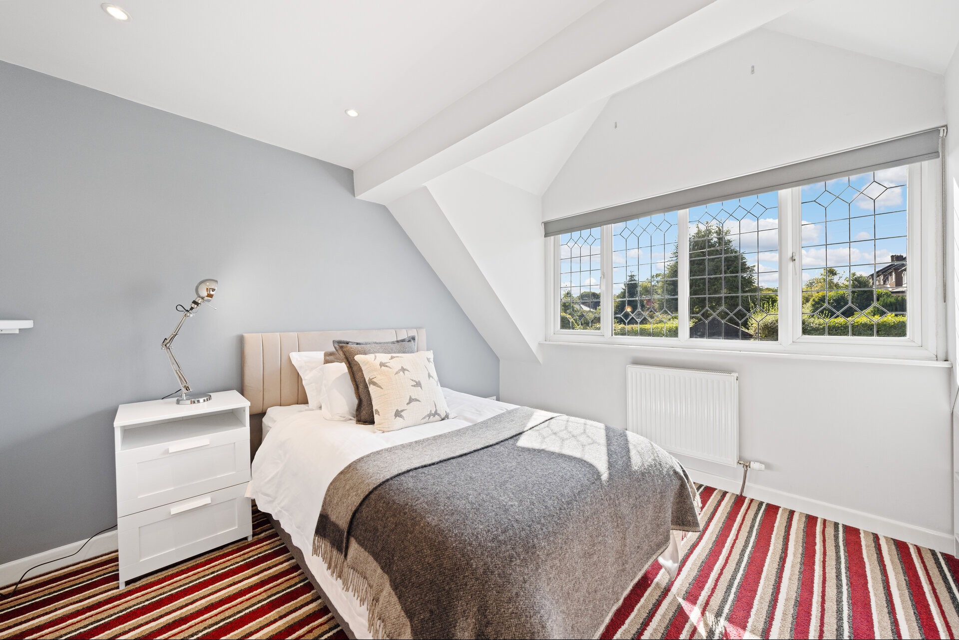 Jasna sypialnia na poddaszu z łóżkiem, stolikiem nocnym i dużym oknem z widokiem na zieleń. Czerwono-biały dywan dodaje wnętrzu charakteru.