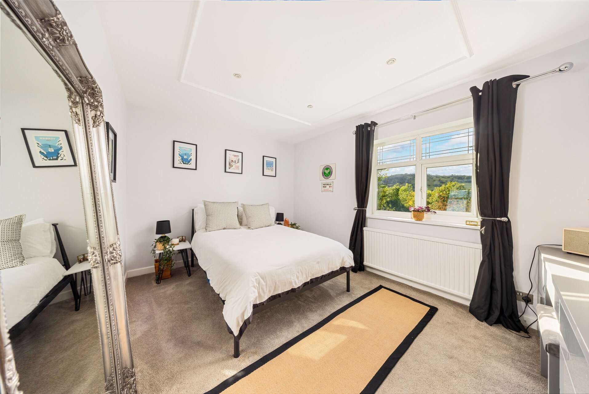 Jasna sypialnia z łóżkiem, dywanem i dużym lustrem w ozdobnej ramie. Widok na zieleń za oknem. Minimalistyczny wystrój z plakatami na ścianie.