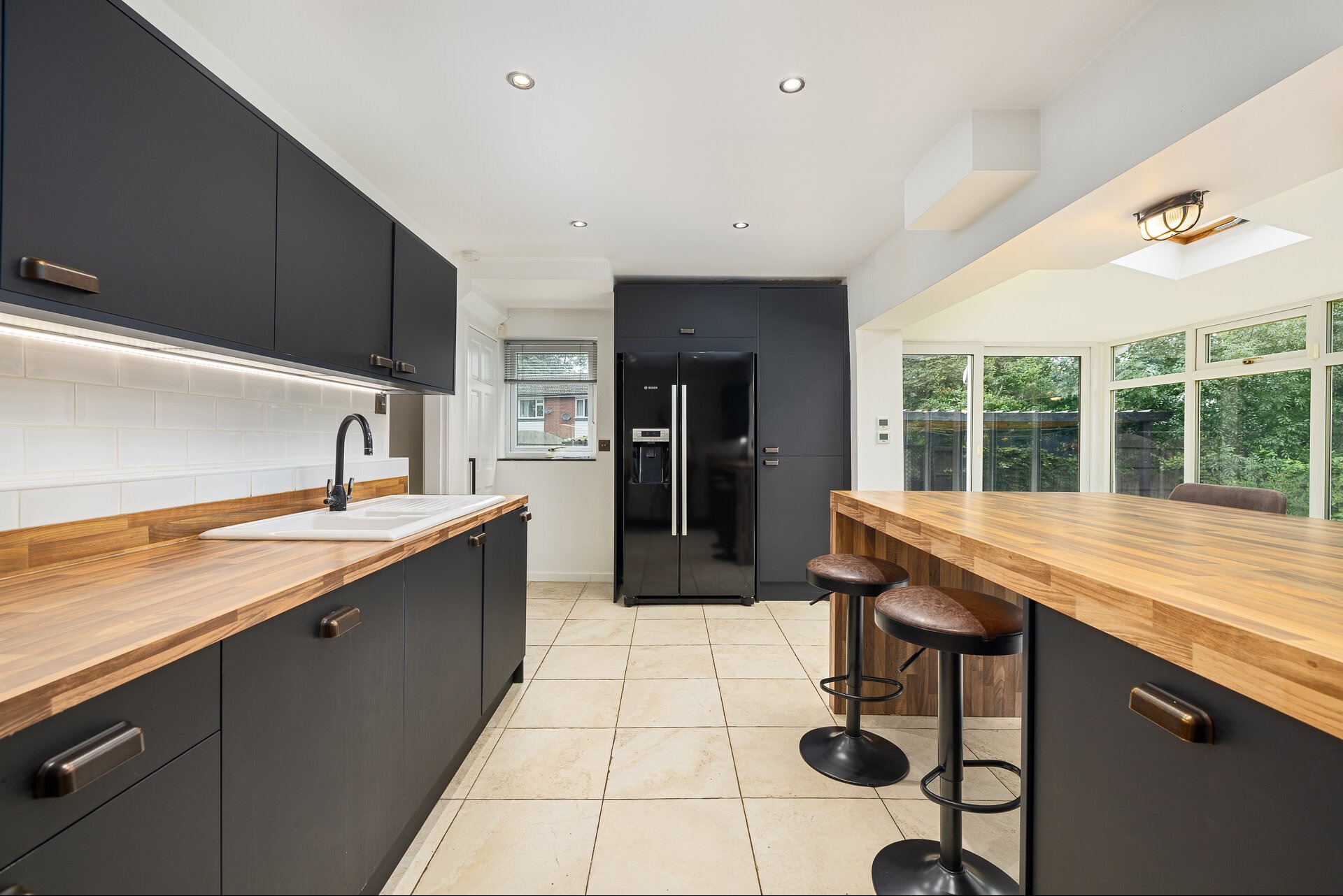 Nowoczesna kuchnia w odcieniach szarości z drewnianymi blatami i wyspą. Uwagę przykuwa czarna lodówka Bosch i designerskie hokery przy wyspie.