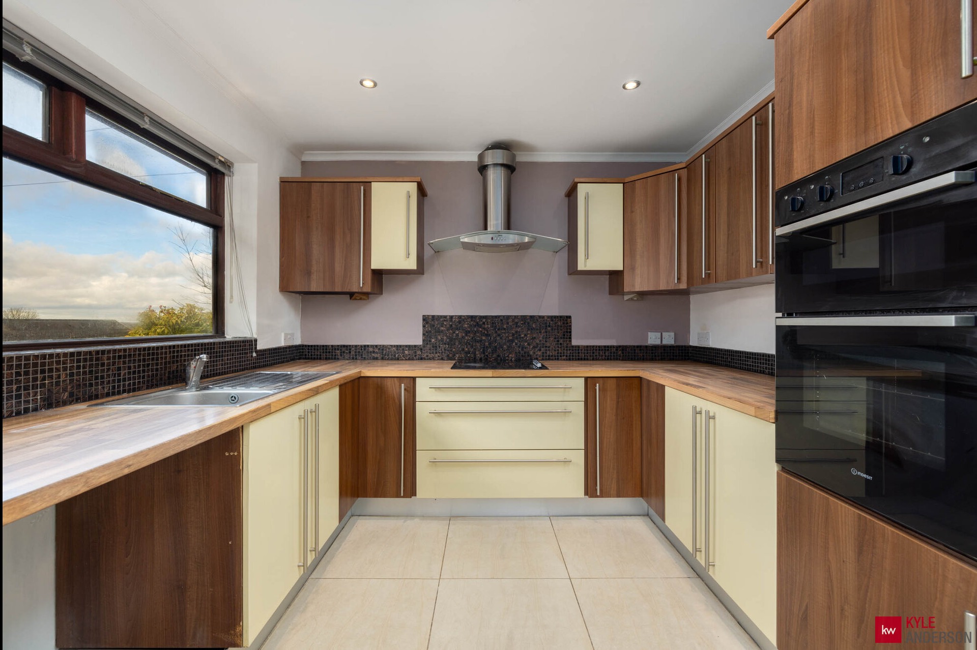 U-kształtna kuchnia z drewnianymi blatami i kremowymi frontami szafek, okno z widokiem na zewnątrz, nowoczesny okap i piekarnik w zabudowie.