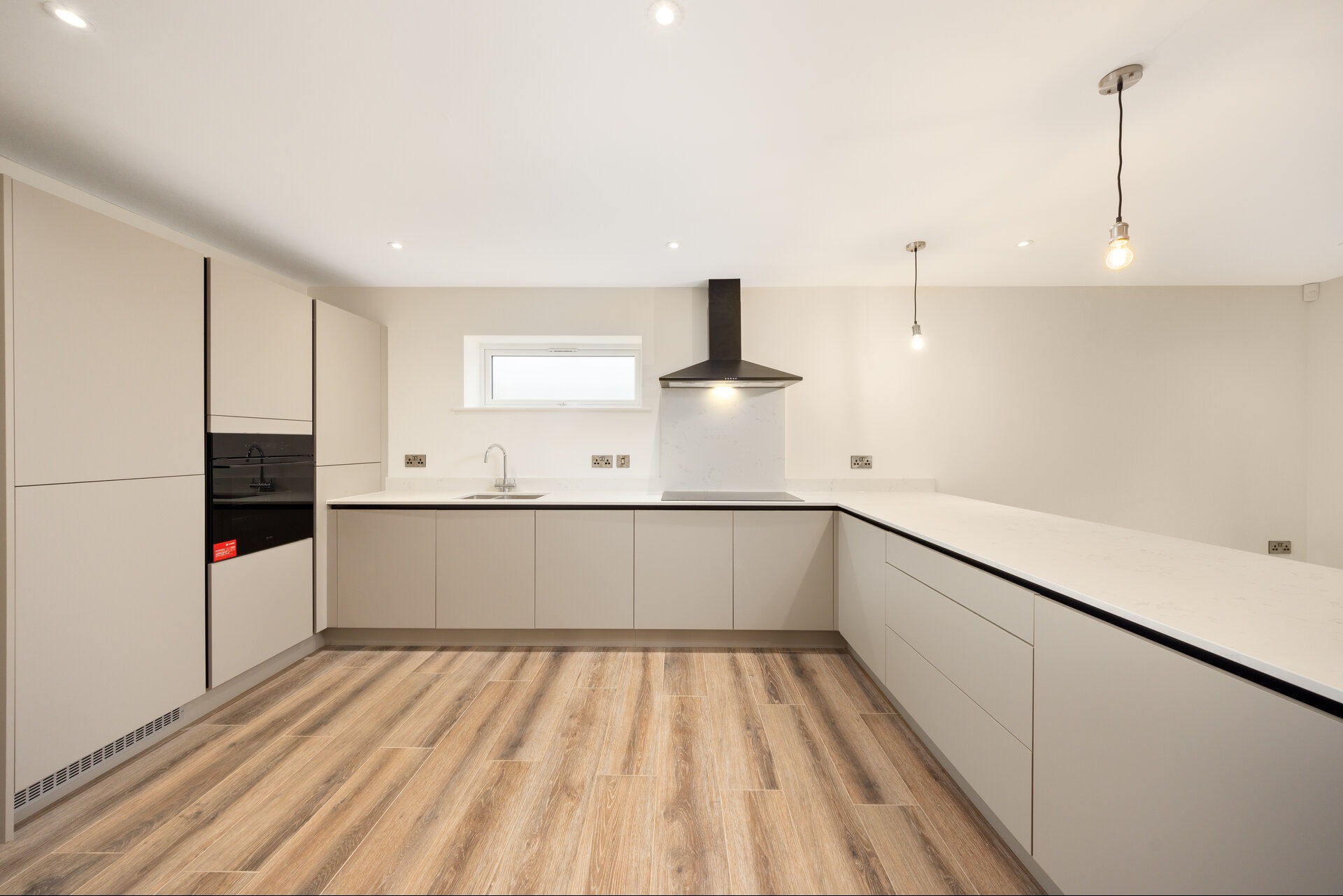 Jasna, nowoczesna kuchnia z drewnianą podłogą, beżowymi szafkami i czarnym okapem. Minimalistyczny design z wiszącymi lampami i białym blatem.