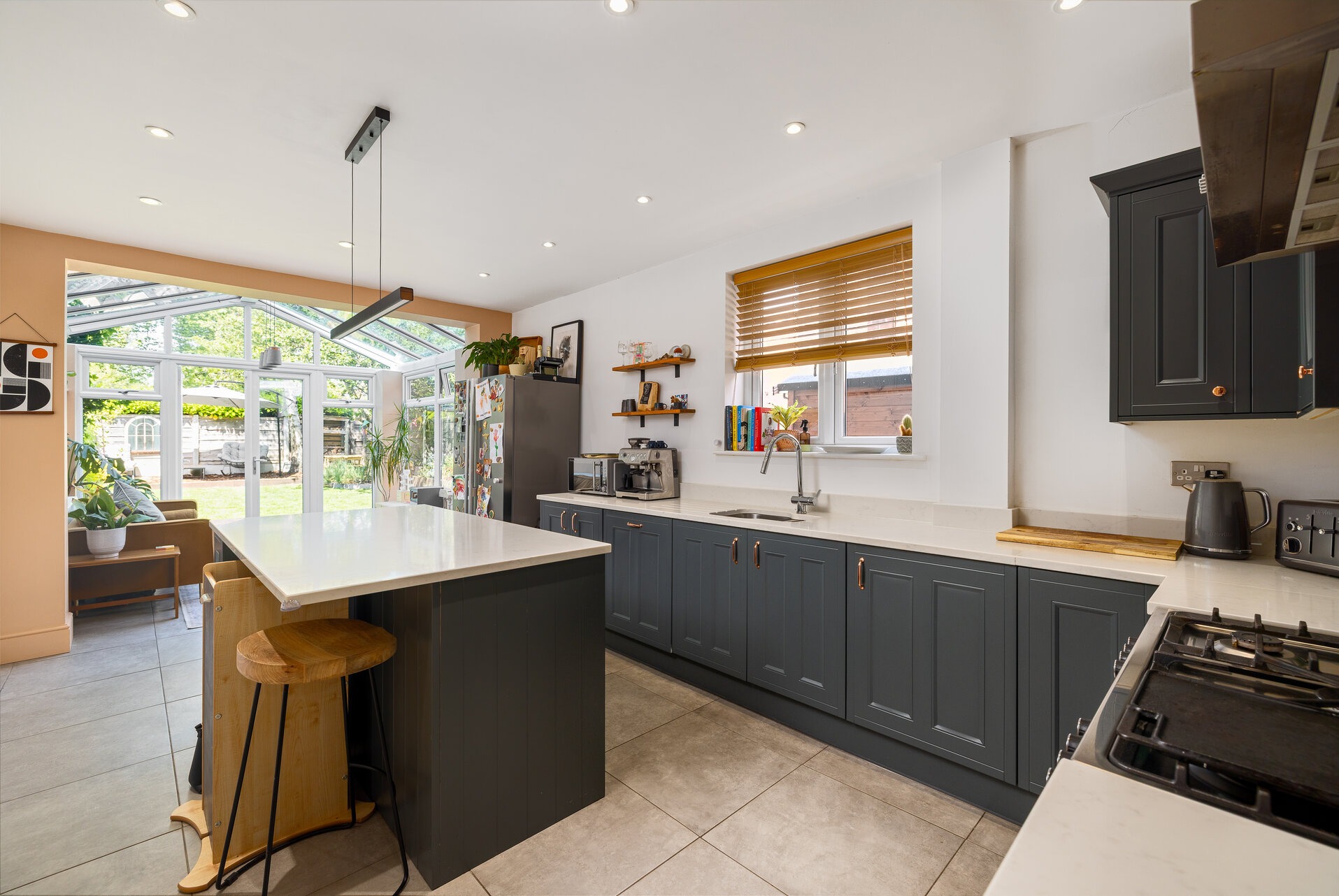 Nowoczesna kuchnia w odcieniach szarości z wyspą i drewnianymi akcentami. Widok na przeszkloną oranżerię z ogrodem w tle. Jasne, naturalne światło.