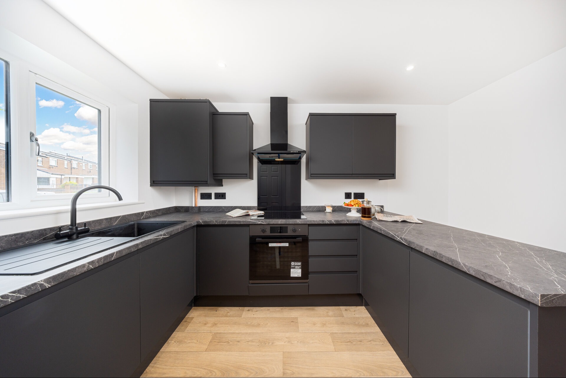 Nowoczesna, minimalistyczna kuchnia w odcieniach szarości z drewnianą podłogą. Widok na okno z błękitnym niebem. Na blacie o marmurowej fakturze leży gazeta i owoce.