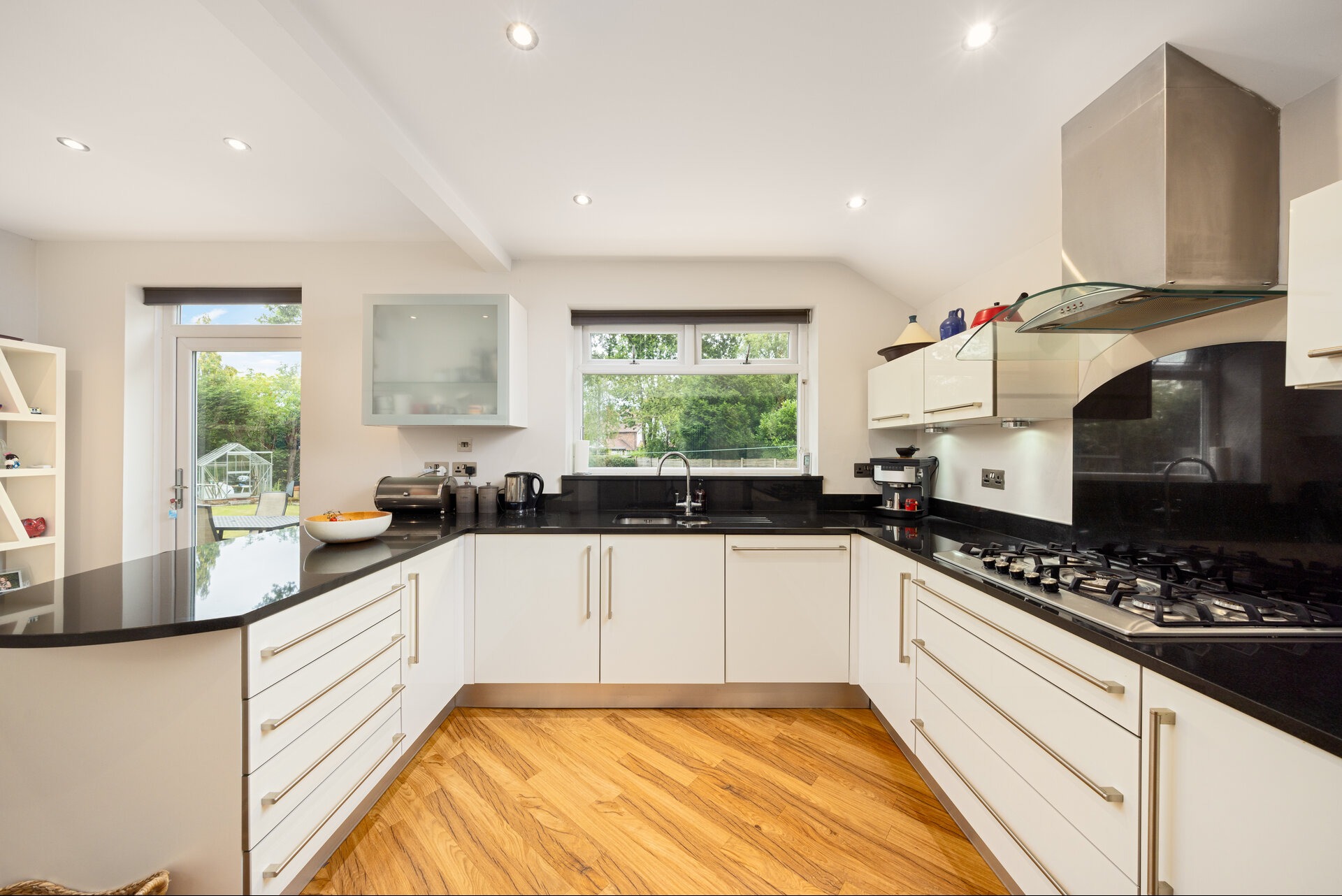 Nowoczesna kuchnia w Warszawie: białe szafki z czarnym blatem, drewniana podłoga, duże okno z widokiem na ogród. Jasne oświetlenie punktowe.