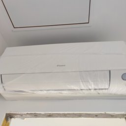 Pawlak Solutions - Biały klimatyzator Daikin, zabezpieczony folią ochronną, zamontowany na ścianie pod sufitem. Widoczny fragment otworu rewizyjnego w suficie i fragment ościeżnicy.