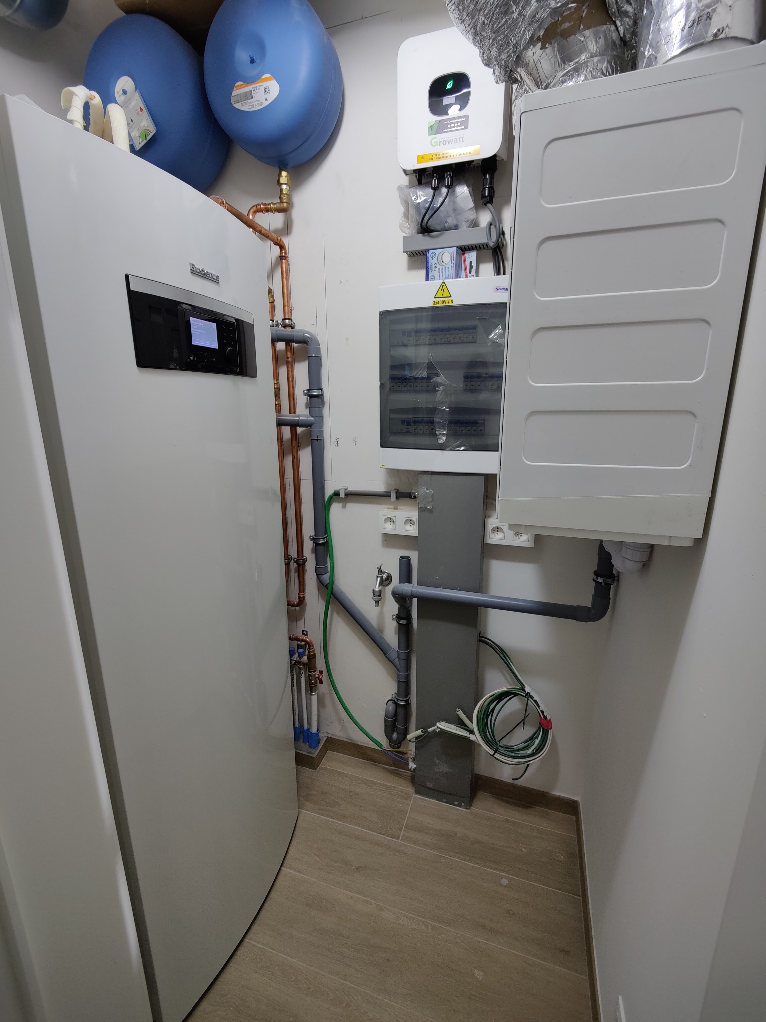 Zmontowana instalacja: pompa ciepła Buderus, inwerter Growatt, zbiorniki wyrównawcze i rozdzielnia elektryczna. Widoczne rury miedziane i PCV, okablowanie. Całość w pomieszczeniu technicznym.