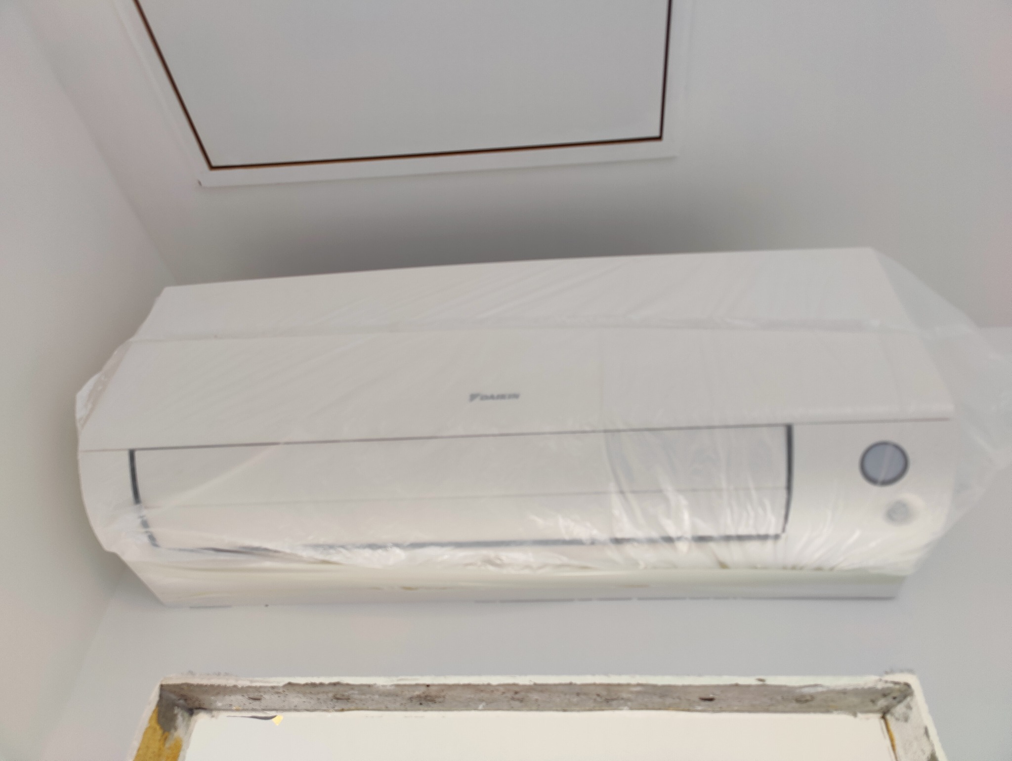 Biały klimatyzator Daikin, zabezpieczony folią ochronną, zamontowany na ścianie pod sufitem. Widoczny fragment otworu rewizyjnego w suficie i fragment ościeżnicy.