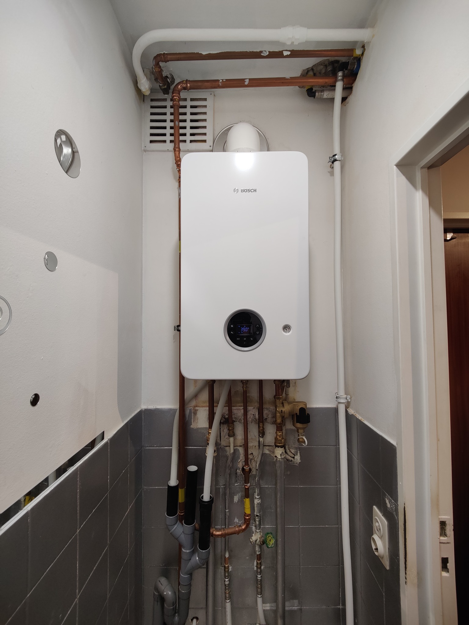 Biały, wiszący piec gazowy Bosch z podłączonymi miedzianymi rurami w małej kotłowni z szarymi płytkami. Widoczne elementy instalacji i wentylacji.