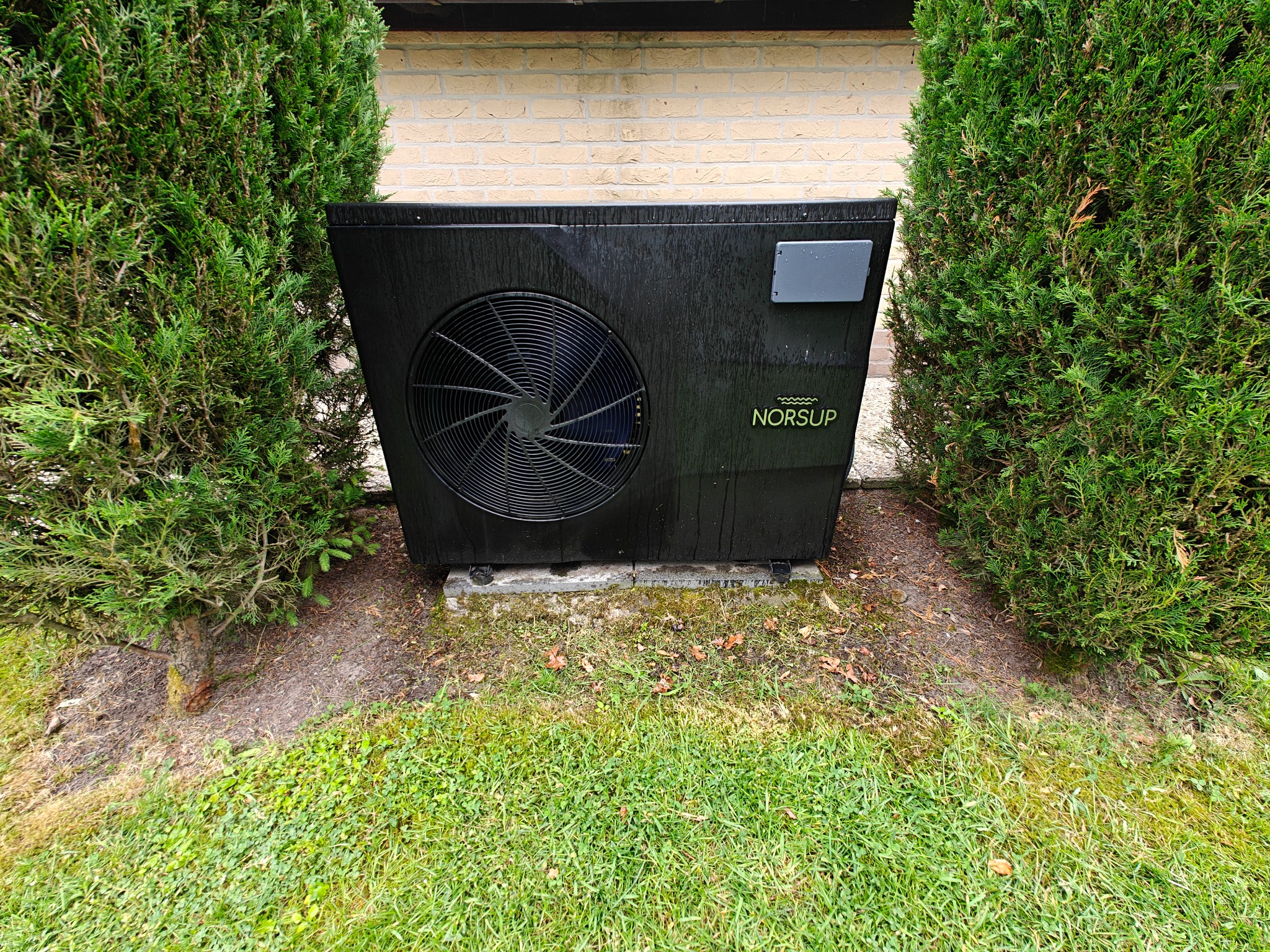 Czarna pompa ciepła Norsup, umieszczona na zewnątrz budynku, otoczona zielonymi krzewami i trawnikiem. Widoczne ślady wilgoci na obudowie.
