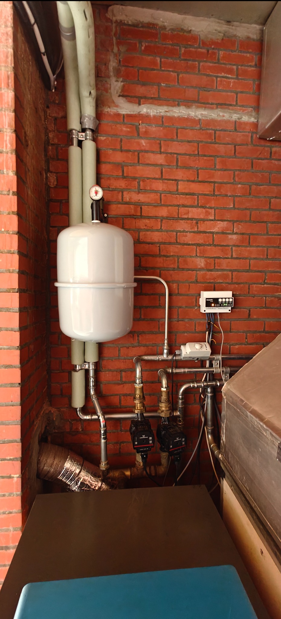 Biały zbiornik wyrównawczy z manometrem, rury i pompy obiegowe na tle ściany z czerwonej cegły. Elementy instalacji grzewczej w kotłowni.
