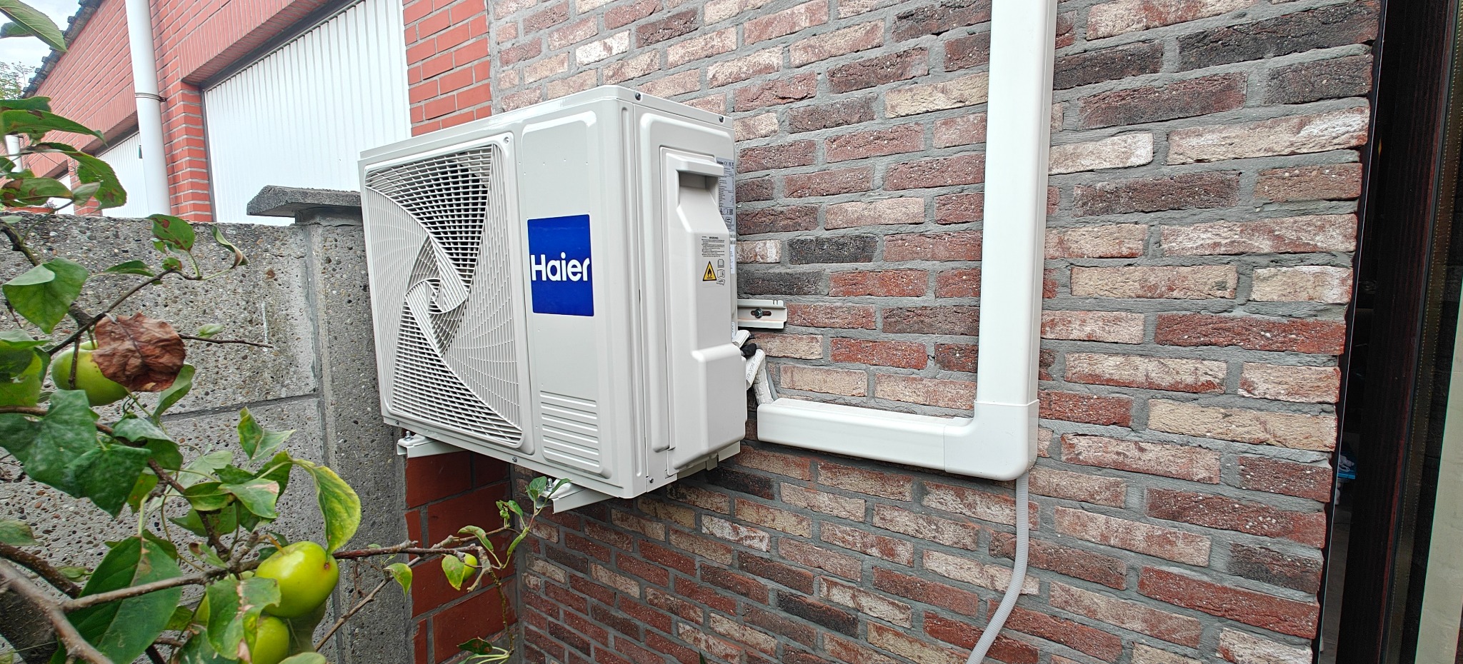 Biała jednostka zewnętrzna klimatyzacji Haier zamontowana na ceglanej ścianie budynku, z widocznymi przewodami i elementami montażowymi. W tle zieleń drzew.