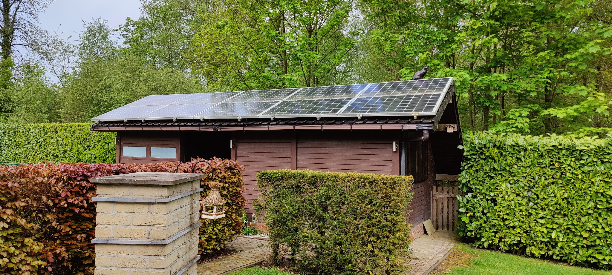 Brązowy budynek gospodarczy z panelami słonecznymi na dachu, otoczony zielenią w Gdańsku. Na dachu siedzi czarny kot.