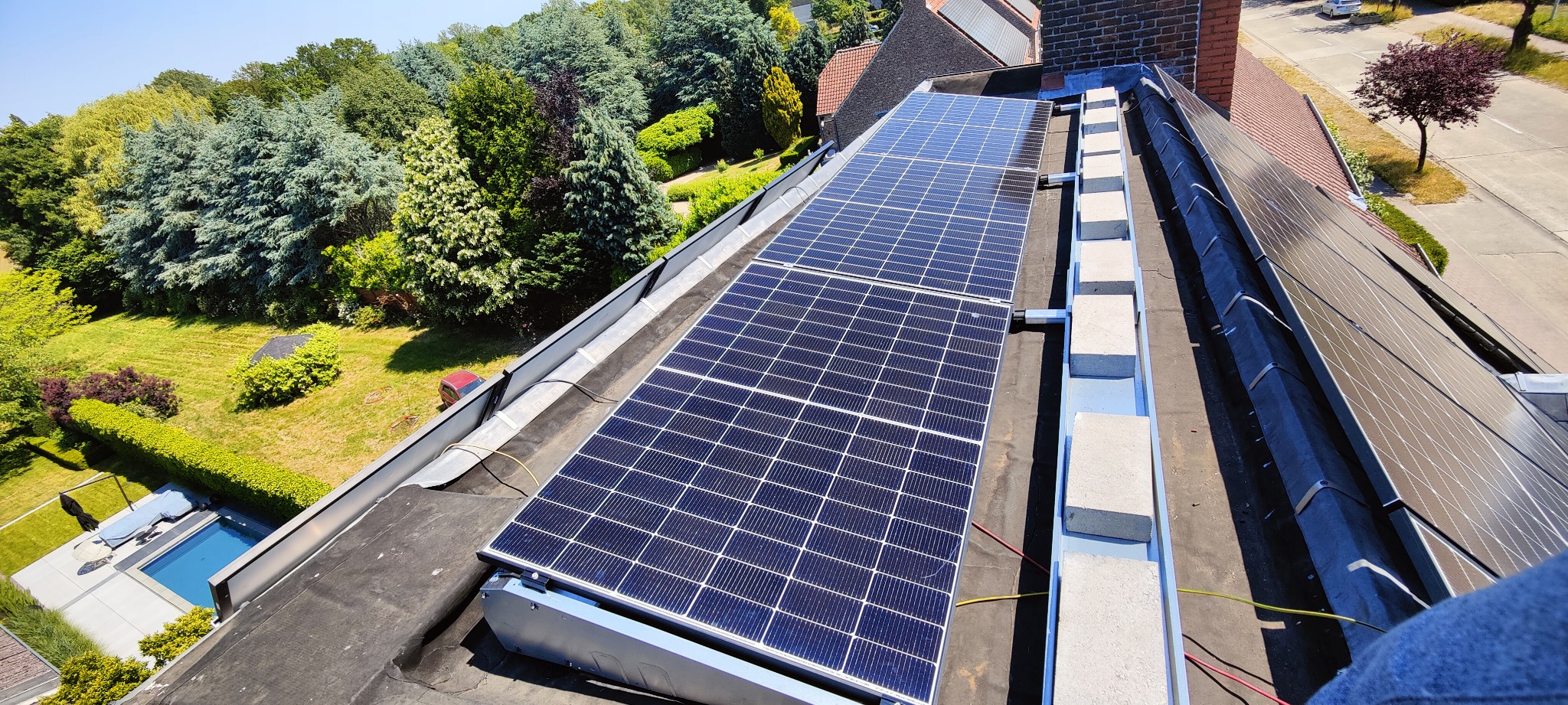 Panele fotowoltaiczne na dachu domu, widok z góry, zieleń w tle. Instalacja solarna z bliska, równomierny układ paneli, elementy montażowe widoczne.