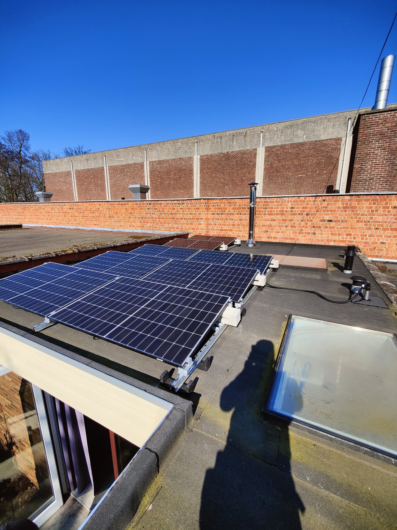 Panele fotowoltaiczne na płaskim dachu budynku z cegły, widoczne elementy montażowe i okno dachowe. Czyste, błękitne niebo w tle. Cień osoby w lewym dolnym rogu.