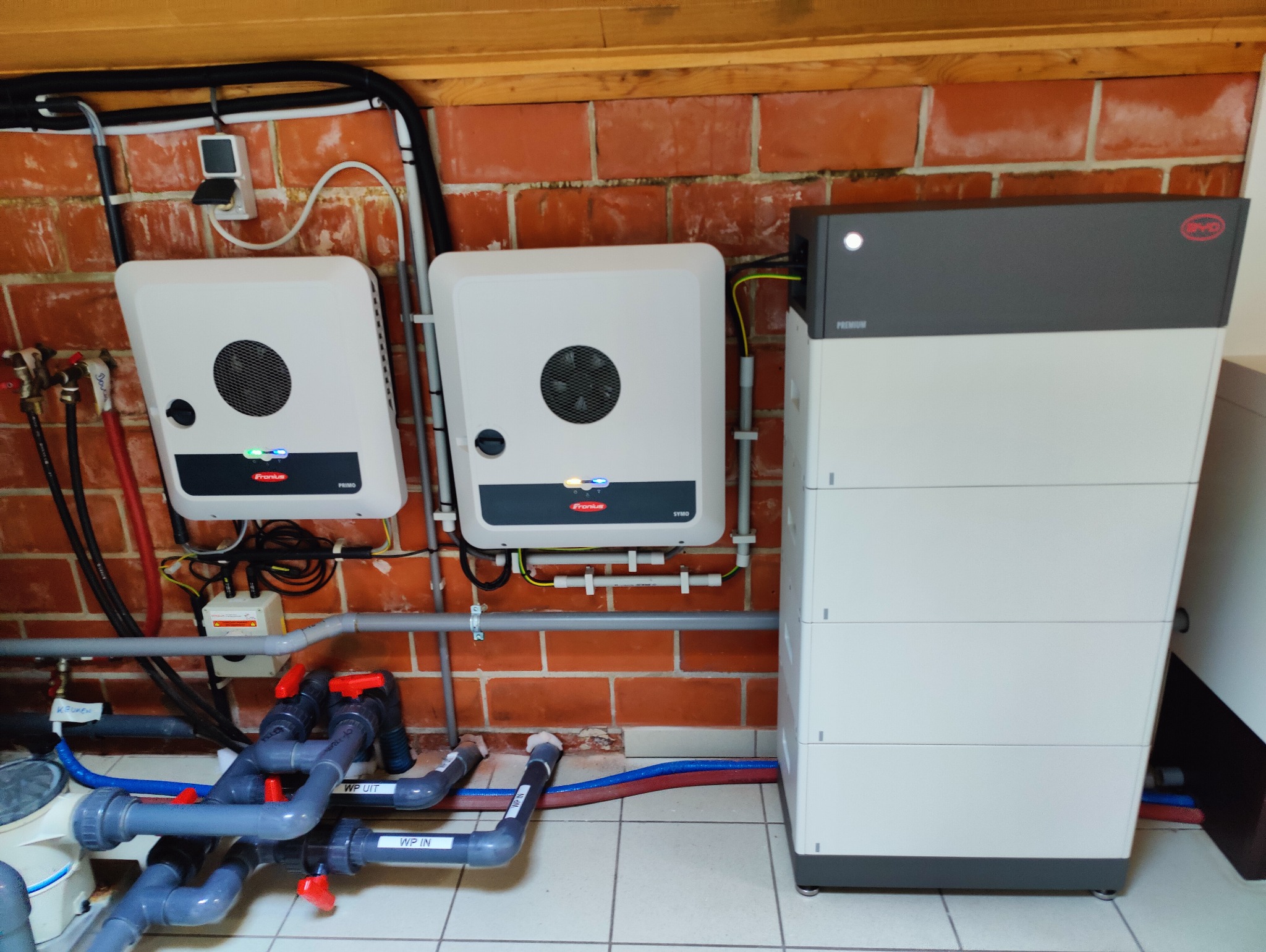 Dwa inwertery Fronius i bateria BYD na tle ceglanej ściany. Widoczne rury instalacji hydraulicznej. Elementy instalacji fotowoltaicznej w pomieszczeniu.