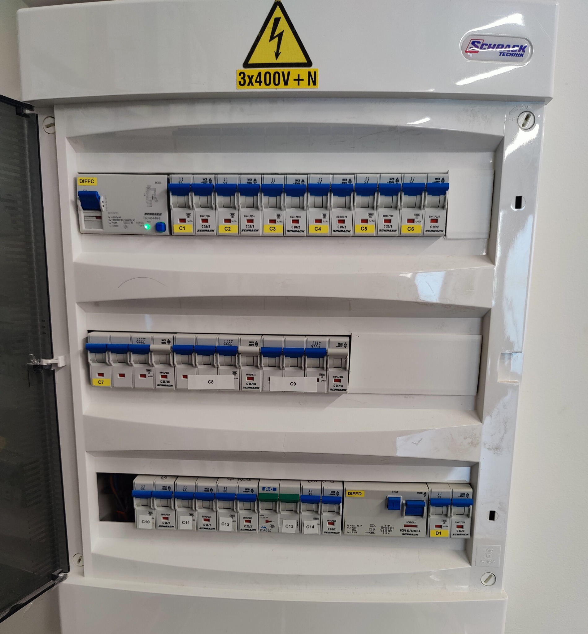 Otwarta skrzynka elektryczna Schrack z bezpiecznikami oznaczonymi C1-C14 i D1, z żółtą naklejką ostrzegawczą 3x400V+N, widok z bliska na instalację elektryczną.