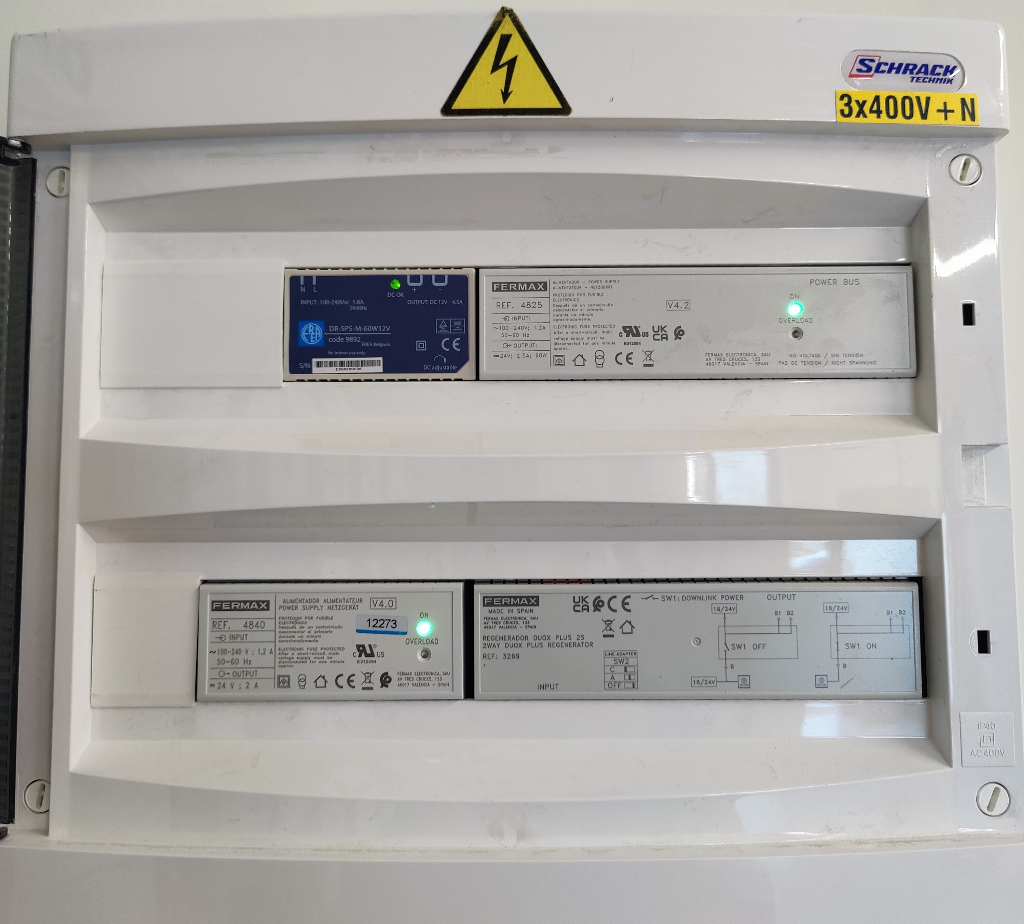 Rozdzielnica elektryczna z modułami zasilania i regeneratorami marki Fermax, widoczne oznaczenia napięć i certyfikaty. Żółty znak ostrzegawczy na górze.