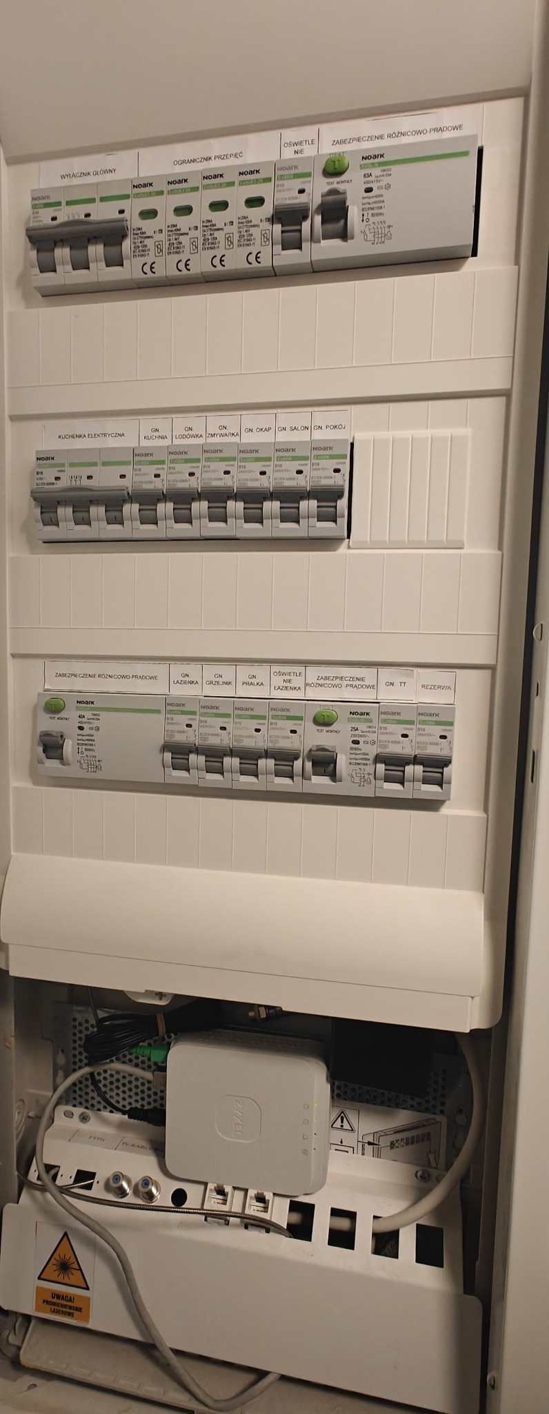 Rozdzielnica elektryczna z bezpiecznikami Noark, opisana dla różnych obwodów (kuchnia, łazienka, oświetlenie). Dodatkowo widoczny router Zyxel i ostrzeżenie laserowe.