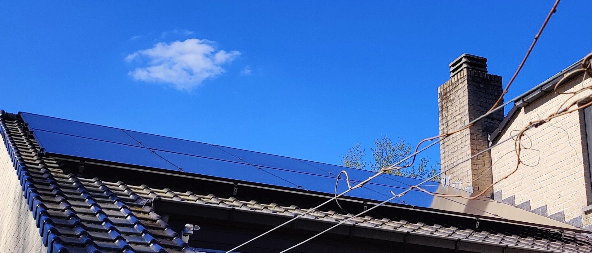 Błyszczące panele fotowoltaiczne na dachu domu z ceglanym kominem na tle błękitnego nieba z chmurą. Widok z dołu, z bliska.
