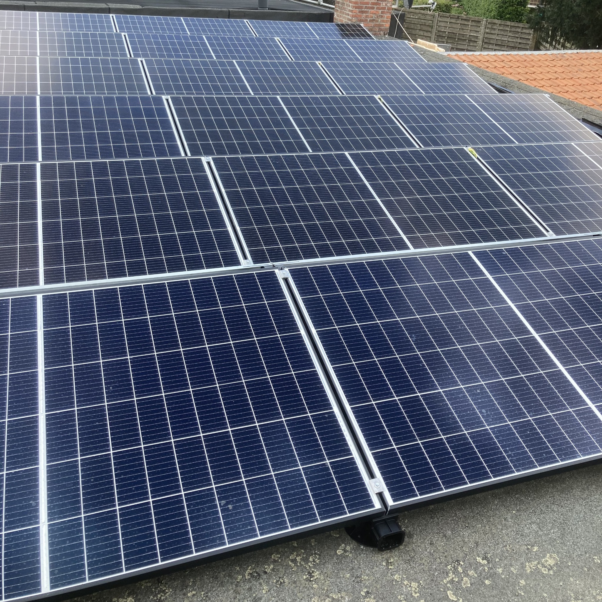 Rząd paneli fotowoltaicznych na dachu, widoczne mocowania i fragment dachu sąsiedniego budynku. Układ paneli w regularnej siatce, z wyraźnymi liniami podziału.