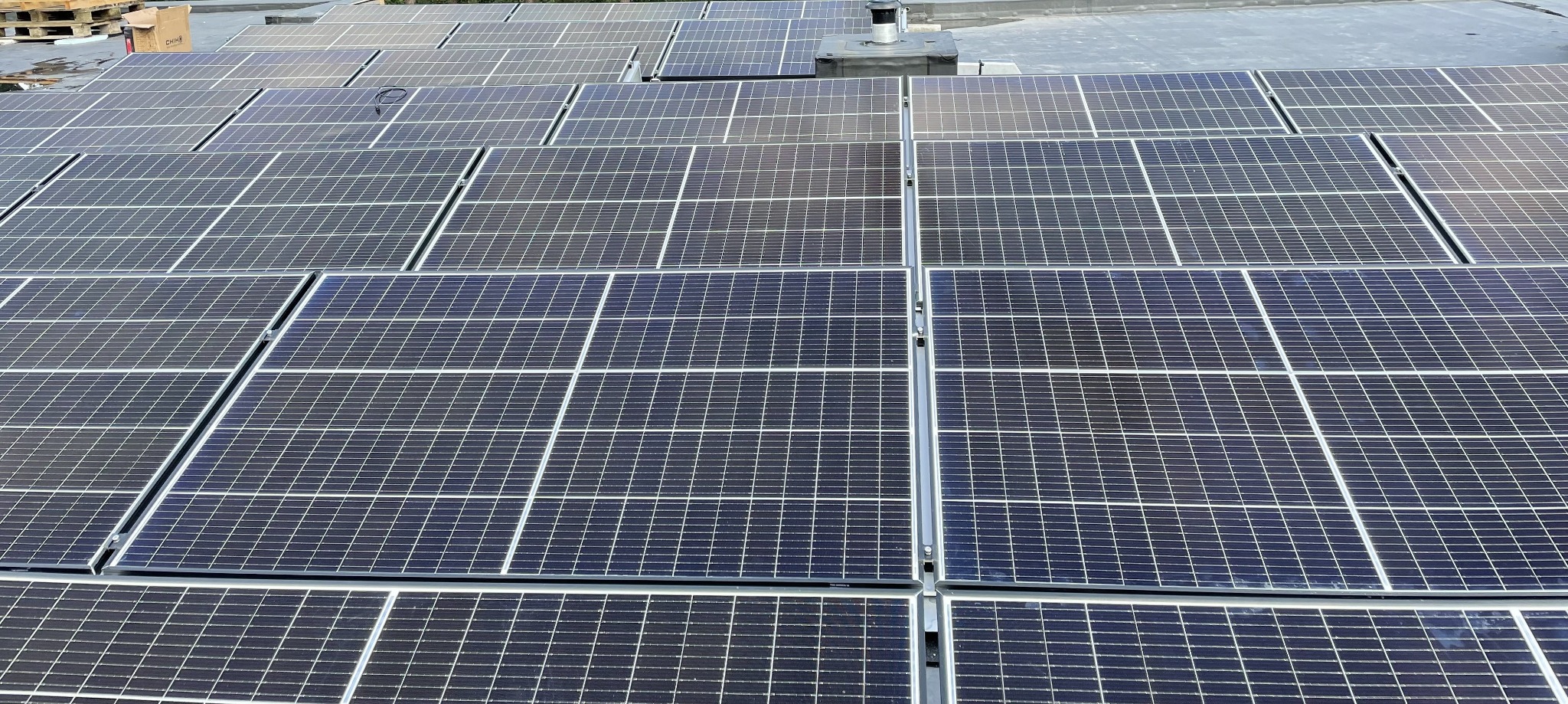 Rząd paneli fotowoltaicznych na płaskim dachu, widok z góry, z widocznym kominem wentylacyjnym w tle. Układ paneli w regularnej siatce, z metalowymi ramami.