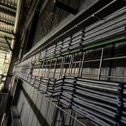 ELEKTRYKA NORBERT SIEKAŃSKI ELENS - Ułożone równolegle kable w metalowej korycie kablowej, widok z dołu pod kątem. Jeden kabel w kolorze zielonym. Szara ściana w tle.