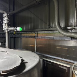 ELEKTRYKA NORBERT SIEKAŃSKI ELENS - Szara instalacja przemysłowa z rurami, zaworami i zielonym czujnikiem na tle metalowej ściany z widocznymi kablami. Elementy instalacji częściowo poza kadrem.