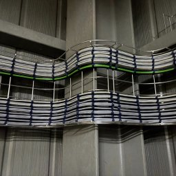 ELEKTRYKA NORBERT SIEKAŃSKI ELENS - Metalowa korytka kablowa z ułożonymi przewodami w szarym pomieszczeniu. Kable spięte niebieskimi opaskami, jeden kabel w kolorze zielonym. Widok z dołu na instalację.