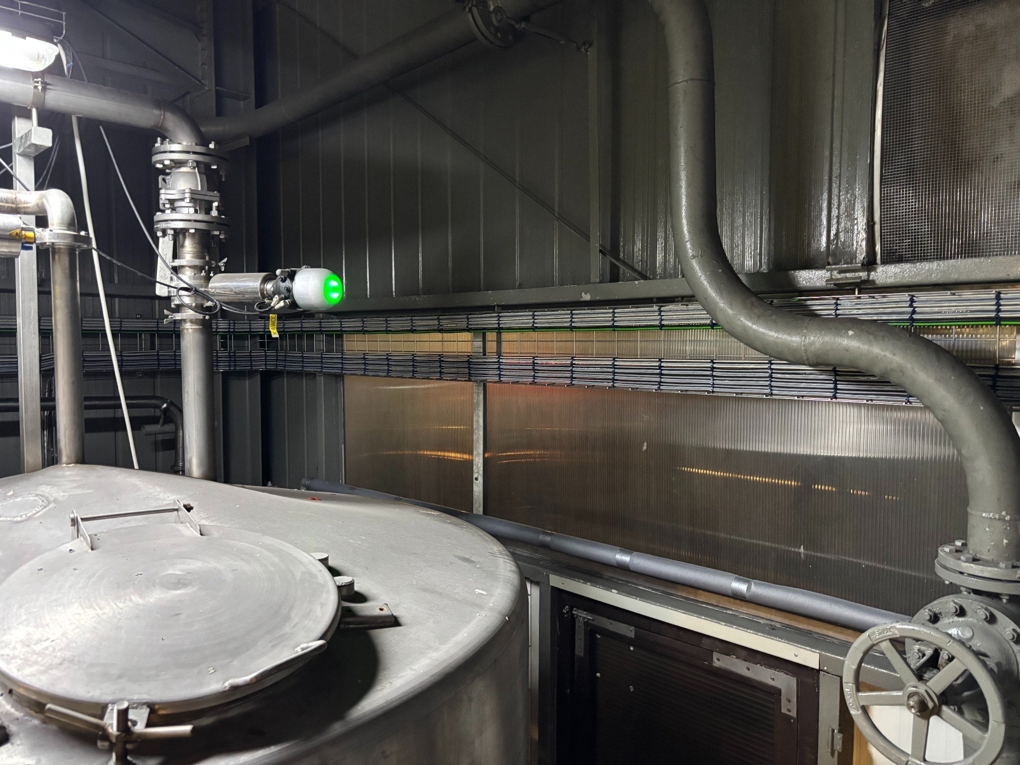 Szara instalacja przemysłowa z rurami, zaworami i zielonym czujnikiem na tle metalowej ściany z widocznymi kablami. Elementy instalacji częściowo poza kadrem.