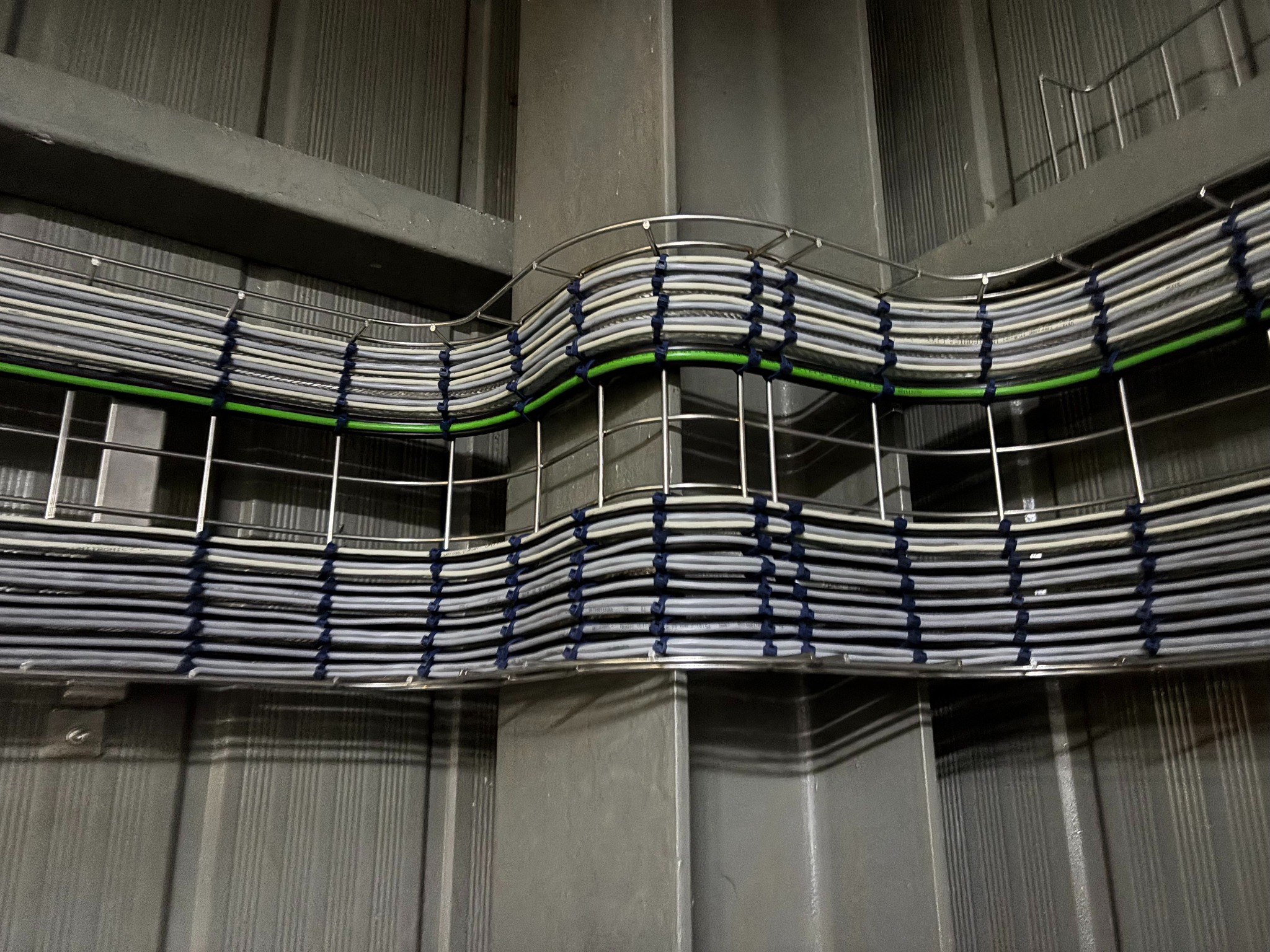 Metalowa korytka kablowa z ułożonymi przewodami w szarym pomieszczeniu. Kable spięte niebieskimi opaskami, jeden kabel w kolorze zielonym. Widok z dołu na instalację.