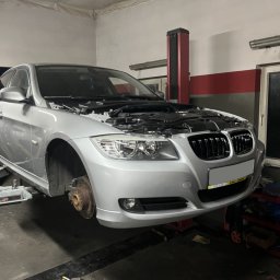 WW-CARS Naprawa Pojazdów Wiesław Wawrzyniak - Srebrny samochód na podnośniku w warsztacie, z otwartą maską i zdjętym kołem, ukazujący etap naprawy. Widoczne narzędzia i logo warsztatu w rogu.